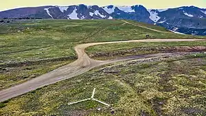 An aerial view of the emergency backcountry helicopter landing zone located at the summit of Rollins Pass; the white prong of the landing zone, furthest from the camera, points north. The comparatively low saddle of Rollins Pass is visible in this image as the summit itself (in the foreground and midground) is plainly lower in elevation than the surrounding mountains. The wood debris consists of both remnants from snowsheds that covered the tracks as well as discarded railroad ties that were removed from service in the summer of 1936.