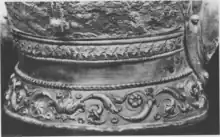 Black and white photograph of the neck guard of the Emesa helmet, showing the three layers of decoration: a torus of ivy leaves bordered by cords at the top, a smooth silver section in the middle, and an acanthus scroll decoration at the bottom.