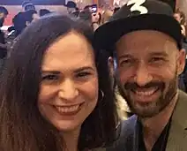 A 39-year-old woman with brown hair and a man with a dark beard and a cap smiling at the camera.