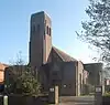 A small brown brick structure unrelieved by dressings or other ornamentation, and partly obscured by shadow, trees and a noticeboard. In the centre is a relatively tall, narrow tower with two arched openings near the top.