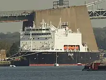 A large ship with a black hull and white superstructure.  Four orange lifeboats are visible midships.  Written on the bow is "EMPIRE STATE".  Several small sailboats are moored in front of it, and the Throgg's Neck Bridge is visible behind.