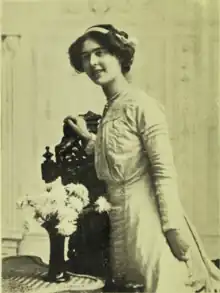 B&W portrait photo of a young woman in a pale dress standing by a table which has flowers in a vase.