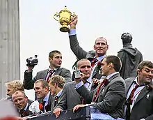 Photograph of nine members of the England rugby team on an open top bus victory parade. Lawrence Dallaglio is in the centre holding up the golden coloured Webb Ellis Cup, which is the trophy awarded to the winners.