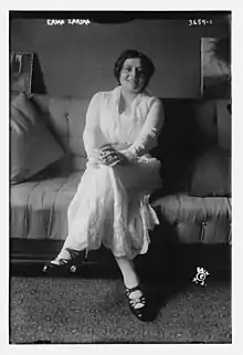 A young woman, seated on a bed and smiling, wearing a white dress and black heeled shoes.