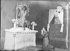 Black and white photo showing a young woman kneeling, praying before an altar with a statue of the Virgin Mary.