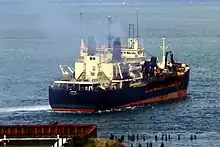 A large, almost rectangular black ship with a red hull creates a mild wake as it moves away from the shore into a very large river. Smoke rises above its decks, which are cluttered with antennas, mechanical fittings, and what appear to be self-contained rooms with multiple windows.