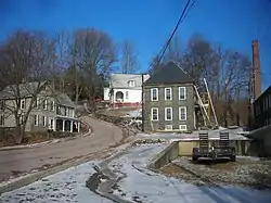 An image showing a curved road, with a single beige victorian house on its left side and three houses, one of bluestone and two others of white clapboard, on the road's right side. To the extreme right of the image is the side of a bluestone factory with a prominent smokestack.