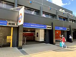 Passengers cross a paved concourse towards escalators that head down into the station entrance. A large "Underground" sign is suspended from a pole above the entrance.