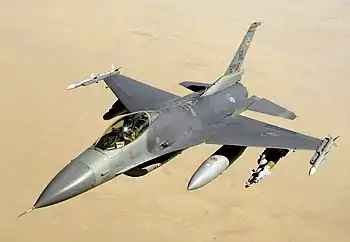 Aerial view of jet aircraft, carrying cylindrical fuel tanks and ordnance, overflying desert