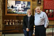 A caucasian known as Jenny Murphy in jeans, a navy long sleeve, a white collared undershirt, and a black blazer is standing next to Jimmy Russel, a taller caucasian man who is bald on top, wearing black pants and a light blue button-up shirt. Both individuals are standing before history memorabilia at the Wild Turkey Distillery, including several bottles and older photographs.