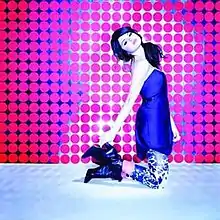 A young woman wearing a blue shirt kneels and poses during a photo-shoot, wearing zebra-printed pants, and high-top boots. The floor is metallic, and the background is filled with several small pink circles.