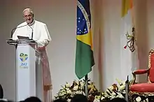 Pope Francis stands at a podium on the left side of the image. On the right are the Brazilian and Vatican flags