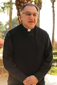 Man in a black cassock with a clerical collar standing with his clasped-hands hanging in front. He has a gentle expression on his face.