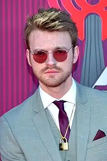 A red-haired man wearing glasses, white shirt, gray suit and purple tie while staring at the camera.