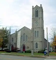 A light gray stone building seen from across a street with a pointed roof in the middle, a tower on the left and two sets of pointed-arch red doors in the center and right. A banner on the left of the center door says "Celebration" in vertical type