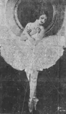 A young white ballerina with bobbed dark hair, wearing a tutu and en pointe, with arms crossed against her chest