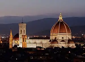 The Cathedral of Saint Mary of the Flower, illuminated at night, showing the large red brick dome, a decorated white marble nave, and a vertical, white marble bell tower to the left. Mountains are visible in the background and a dark, low-lying city in the foreground.