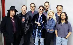 Seven men and one woman gathered together in front a photographic sheet, smiling at the photographer (Jay Blakesberg), wishing they were backstage at a concert about to start.