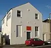 Three-quarter view of a tall, two-storey, plain-walled, white-painted building with a pointed roof. The front has one blank window and a half-blank, half-glazed window flanking a narrow door, above which is an orange sign.  There are also six circular protrusions.  The side wall has three windows and a thin metal flue.