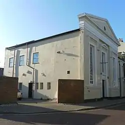 Three-quarter view of a wide, long stuccoed building, with the side wall more prominent. This is mostly a blank wall, but there are three windows towards the rear.  The façade has three tall windows tapering from bottom to top; the central window has a small pediment and is shorter than the others to accommodate an entrance door.  Above the roofline is a large pediment with louvred circular opening.