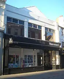 A wide, flat-roofed shop with a pediment. The lower storey has a black shopfront with plate glass windows; the upper storey has three wide rectangular windows.  All exposed areas of wall are painted white.