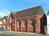 A dark red brick hall on a corner site, with adjoining houses in an identical style. The hall, partly obscured by a traffic light, has a pointed-arched entrance with a wooden door below a rectangular window frame with three lancets.  To the right of the door, there are three plain windows between brick buttresses.  Above the entrance is a steep roof perpendicular to the main roof.