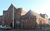 A wide terracotta-coloured brick building in two parts: to the left, a two-storey structure with three lancet windows above the arched entrance porch, spirelets and recessed parts with paired and single lancets, all topped with a triangular roof with vertical recessed sections; and to its right, a rounded single-storey section with single lancets separated by buttresses.