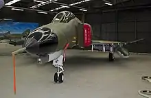 Colour photo of a military jet fighter painted in a camouflage pattern parked inside a black-walled building