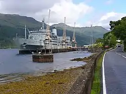 Grey warship docked at a jetty, with hills in the background