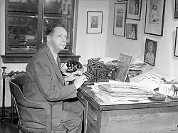 Black and white photo of Hewitt sitting at a desk piled with papers