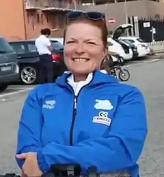 Headshot of a white woman in a bright blue kit with cars in the background.