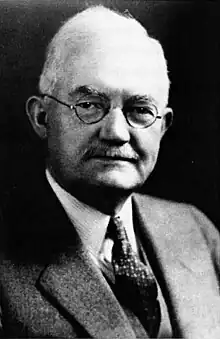 Black and white portrait photograph of an elderly man from the chest up. He wears a suit and glasses with round frames, and he has short, white hair.