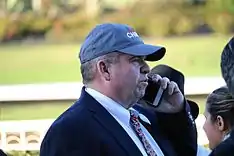 a middle aged man in a dark suit and ball cap, talking on a cell phone