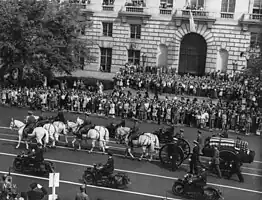 A black-and-white photograph of a wagon being drawn and accompanied by seven white horses, some of which are being ridden by people