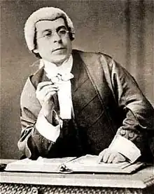  An old photo of an actor in the costume of a judge, with robe and peri-wig, who leans on the bench with a whimsical expression.