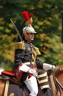 Buste d'un homme à cheval en uniforme noir, rouge et or, le sabre à la main, et coiffé d'un casque doré orné de plumeaux rouges et d'une longue que de cheval noire.