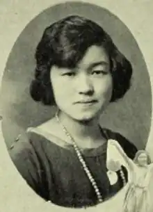 A young Japanese woman with a dimpled chin, with hair dressed to chin length and sideswept bangs, wearing a dark dress or blouse with a wide neckline and a string of beads or pearls, in an oval frame