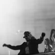 Future with bleached blonde dreadlocks, wearing sunglasses and a black jacket, throws his hands in the air, letting his jacket flow in the wind, while standing in front of a black-and-white foggy background. Three women can be seen standing in the background.