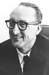 black-and-white image of George Mueller, smiling to the viewer's left in a light shirt, dark tie and suit coat, and wearing dark plastic eyeglasses