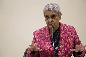 Photograph of an old woman speaking in front of a mic with her both hand in the air.