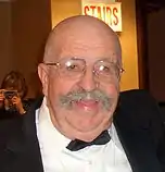 A photograph of a bald man with a grey moustache wearing glasses and a black tuxedo, looking at the camera, and holding a drinking glass