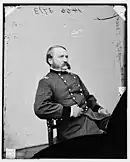 Black and white photo of a seated man with a large moustache and muttonchops. He wears a dark military uniform with two rows of buttons, and the two stars of a major general on the shoulder tabs.