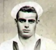 Head of a white man with dark, slicked-back hair wearing a white sailor's cap pushed up on his forehead and a sailor suit with a dark scarf tied around the neck.