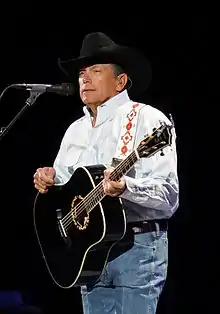 A man wearing a black cowboy hat and a white shirt, playing guitar and singing into a microphone