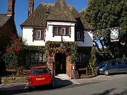 A front view of a two-storey public house, the lower storey in brick and the upper storey plastered.  It has a protruding central bay with a gable, steep tiled roofs and an ornate inn sign protruding diagonally from the right corner.