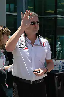 Man in his late thirties, with his right hand elevated. He is wearing a white T-shirt and sunglasses.