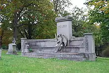 Short, wide memorial lined by a bench with angel statue sitting on the bench. Material is grey and central pillar depicts the name Giles.