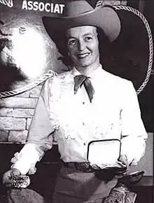 Photograph of a woman dressed in western attire wearing a cowboy hat and holding a trophy belt buckle and trophy box