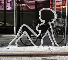 The outline of a silhouette of a naked woman leaning back, in galvanized steel, on a street curb. It has a bicycle with a shopping basket chained to it.