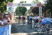 A group of cyclists ride under an arch erected on the road. Spectators watch at the roadside.